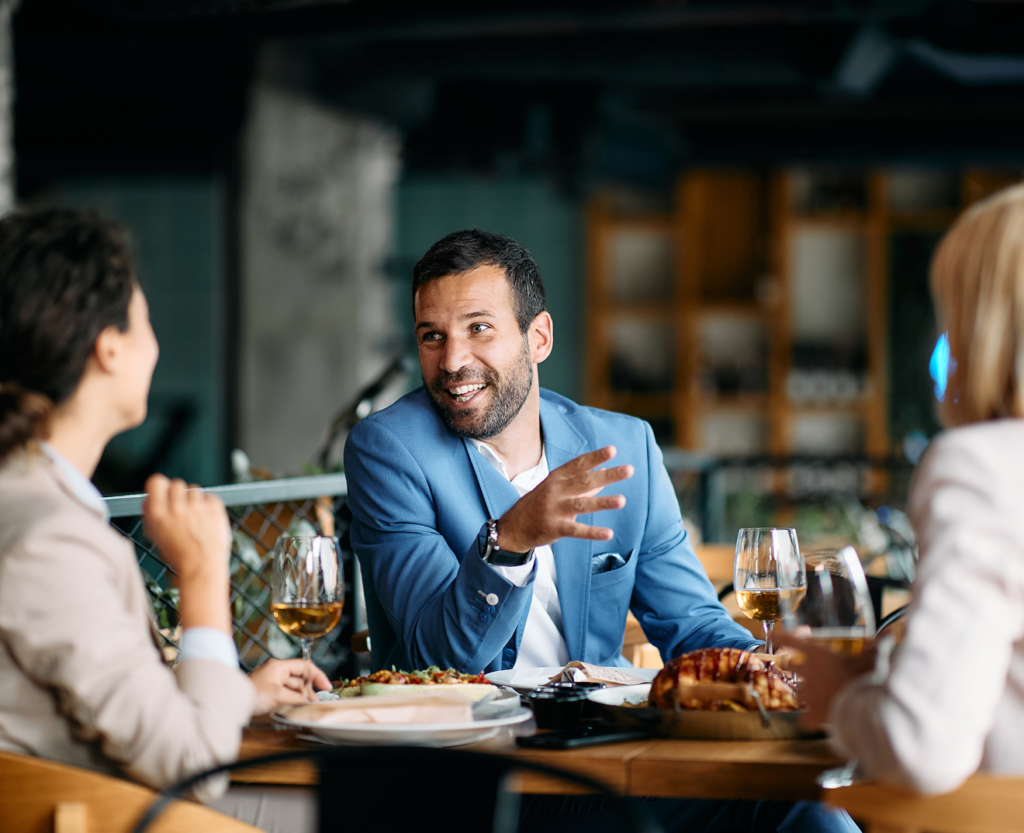 Man talking with two women at a restaurant table with food and drinks.