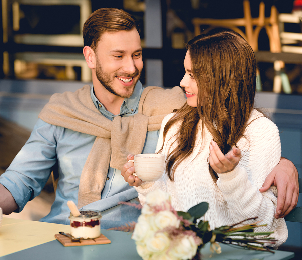 A man and woman smiling at each other at a cafe table with coffee and flowers.
